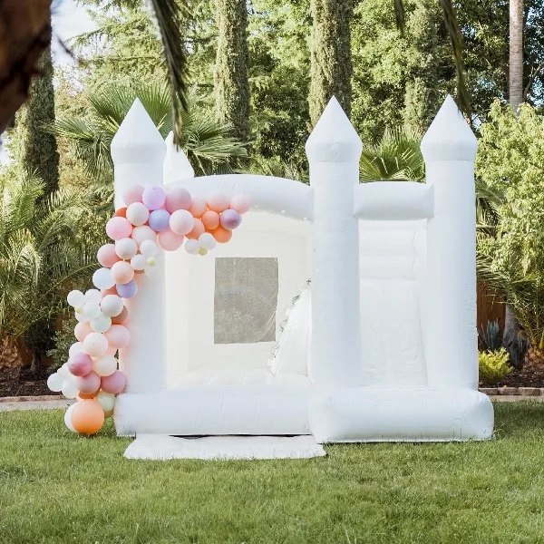 A white inflatable castle backdrop decorated with a pastel pink, orange, and purple balloon garland on a lawn.