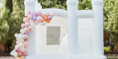 A white inflatable castle backdrop decorated with a pastel pink, orange, and purple balloon garland on a lawn.