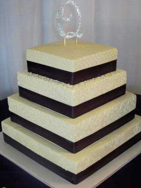 A four-tier square wedding cake with white and dark chocolate bands, textured white icing, and a silver monogram topper.