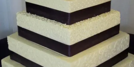 A four-tier square wedding cake with white and dark chocolate bands, textured white icing, and a silver monogram topper.
