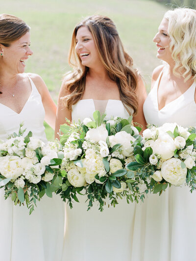 Three women in white dresses holding large white and green floral bouquets outdoors.