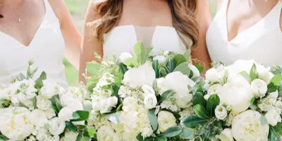 Three women in white dresses holding large white and green floral bouquets outdoors.