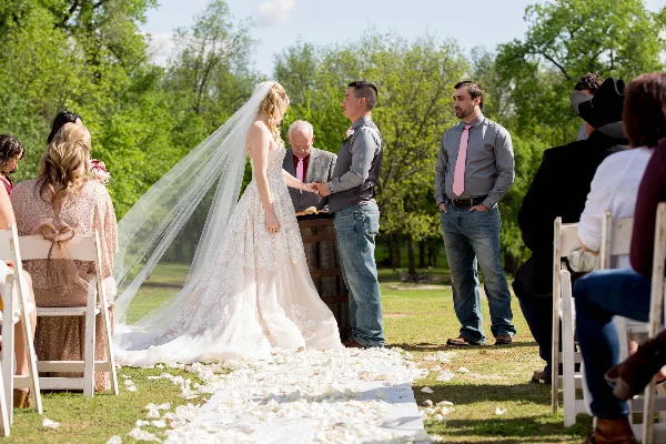 Outdoor wedding ceremony with bride and groom exchanging rings in a grassy area surrounded by trees.