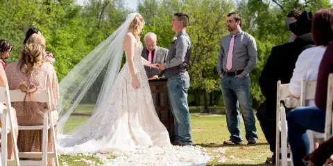 Outdoor wedding ceremony with bride and groom exchanging rings in a grassy area surrounded by trees.