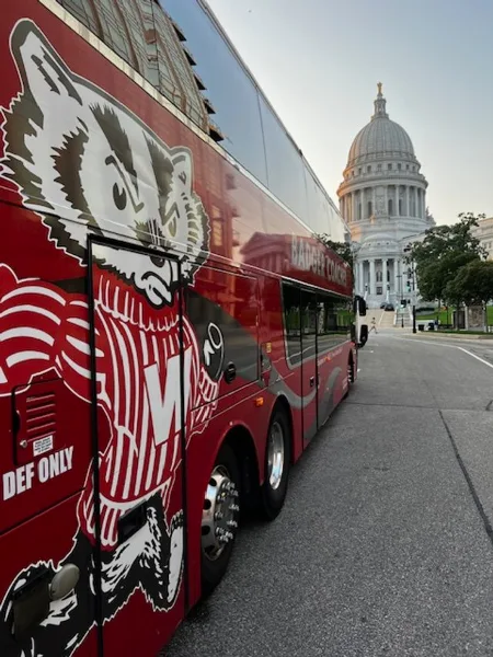 A red Badger Bus motor coach parked on a city street with the Wisconsin State Capitol building in the background.