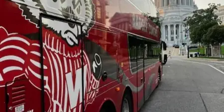 A red Badger Bus motor coach parked on a city street with the Wisconsin State Capitol building in the background.