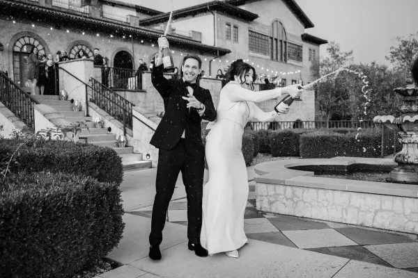 A bride and groom in formal attire popping champagne bottles outdoors at a wedding venue.