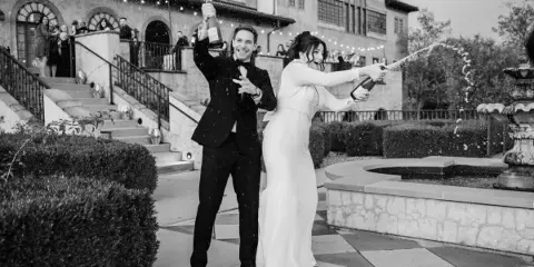 A bride and groom in formal attire popping champagne bottles outdoors at a wedding venue.