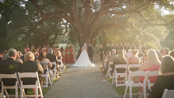 A wedding ceremony taking place outdoors under a large tree with a bride and groom at the altar and guests seated in white chairs.