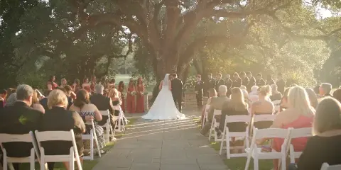 A wedding ceremony taking place outdoors under a large tree with a bride and groom at the altar and guests seated in white chairs.