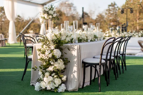 A long wedding reception table set with white linens, tall glass candle holders, and cascading white floral arrangements with greenery.