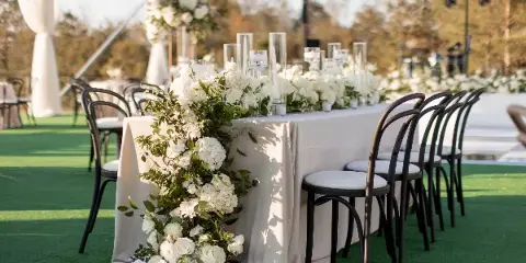 A long wedding reception table set with white linens, tall glass candle holders, and cascading white floral arrangements with greenery.