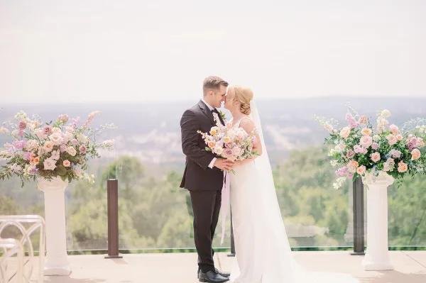 A bride and groom standing face-to-face on a terrace with floral arrangements and a scenic background.