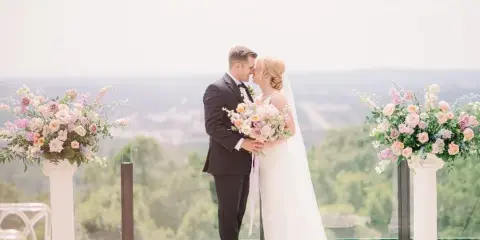 A bride and groom standing face-to-face on a terrace with floral arrangements and a scenic background.