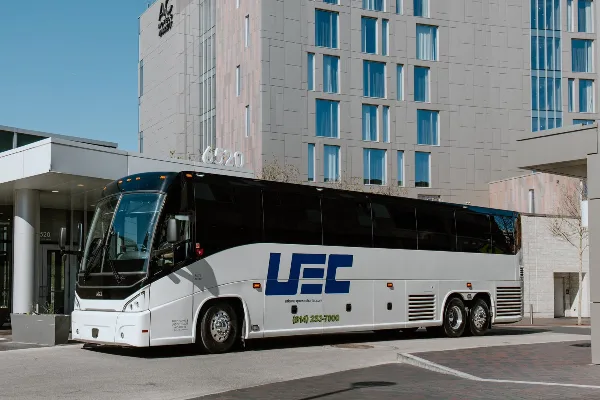 A white and black charter bus with the UFC logo and urbanexpresscharter.com branding parked in front of a modern building.