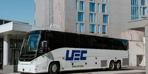 A white and black charter bus with the UFC logo and urbanexpresscharter.com branding parked in front of a modern building.