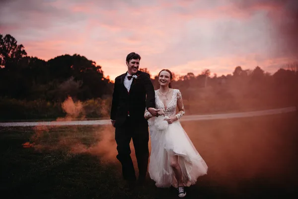 A bride and groom walking hand-in-hand in a field at sunset with smoke effects.