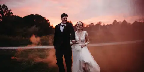 A bride and groom walking hand-in-hand in a field at sunset with smoke effects.
