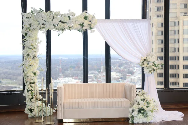 A wedding ceremony setup featuring a white artificial flower arch, white draping, a cream-colored sofa, and floral arrangements in front of large windows overlooking a city.