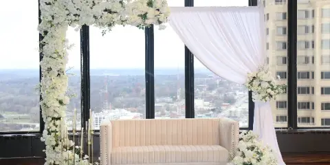 A wedding ceremony setup featuring a white artificial flower arch, white draping, a cream-colored sofa, and floral arrangements in front of large windows overlooking a city.