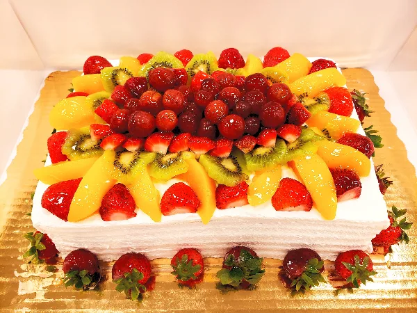 A square white frosted cake topped with an intricate arrangement of fresh strawberries, kiwi slices, mango chunks, and glazed cherries, presented on a gold cake board.