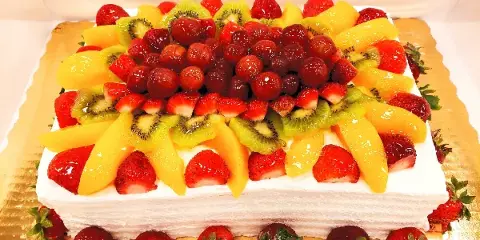 A square white frosted cake topped with an intricate arrangement of fresh strawberries, kiwi slices, mango chunks, and glazed cherries, presented on a gold cake board.
