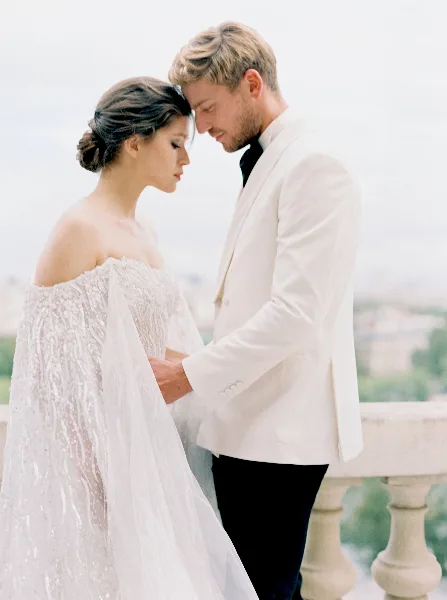 A bride in a white gown and a groom in a white suit standing close together outdoors.