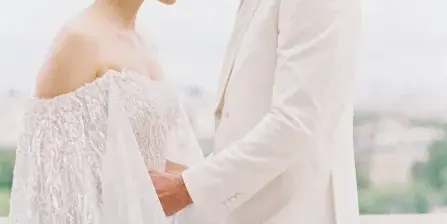 A bride in a white gown and a groom in a white suit standing close together outdoors.