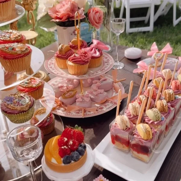 An elegant outdoor dessert table featuring a tiered stand of decorated cupcakes, a plate of pink macarons, a fruit tart, and a row of layered pastry bites topped with macarons.
