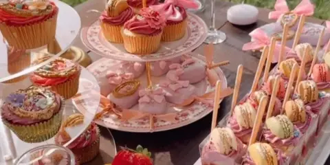 An elegant outdoor dessert table featuring a tiered stand of decorated cupcakes, a plate of pink macarons, a fruit tart, and a row of layered pastry bites topped with macarons.
