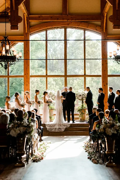 A wedding ceremony in progress inside a rustic venue with large arched windows and wooden beams.