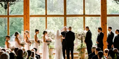 A wedding ceremony in progress inside a rustic venue with large arched windows and wooden beams.