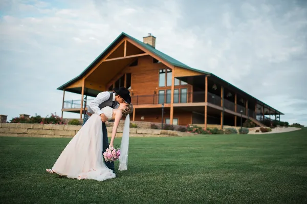 A bride and groom kissing on a lawn in front of a large wooden lodge with a green roof.