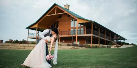 A bride and groom kissing on a lawn in front of a large wooden lodge with a green roof.