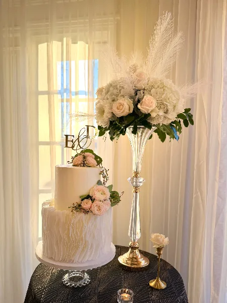 A two-tier white wedding cake decorated with pink roses and a gold 'E & D' topper, displayed next to a tall floral arrangement.