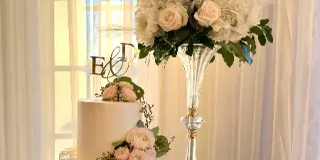 A two-tier white wedding cake decorated with pink roses and a gold 'E & D' topper, displayed next to a tall floral arrangement.