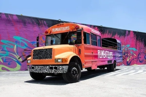 An orange and purple open-air party bus with 'RUMBATOURS' branding parked in front of a colorful graffiti wall.