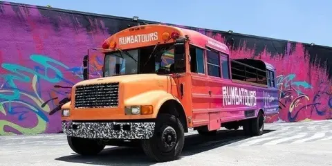 An orange and purple open-air party bus with 'RUMBATOURS' branding parked in front of a colorful graffiti wall.