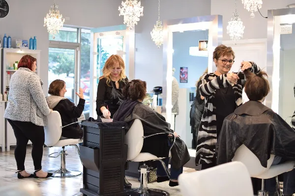 A busy hair salon interior with multiple stylists working on clients in white chairs under crystal chandeliers.