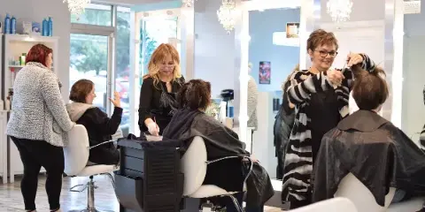 A busy hair salon interior with multiple stylists working on clients in white chairs under crystal chandeliers.