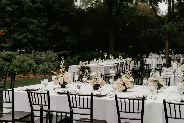 An outdoor wedding reception setup featuring long tables with white linens, black chairs, and floral centerpieces in a garden setting.