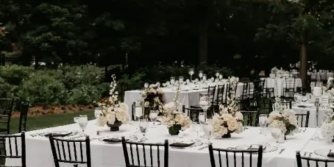 An outdoor wedding reception setup featuring long tables with white linens, black chairs, and floral centerpieces in a garden setting.