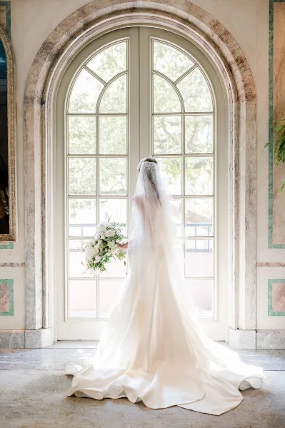 A bride in a white wedding dress and veil stands facing a large arched window, holding a bouquet of white flowers.
