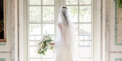 A bride in a white wedding dress and veil stands facing a large arched window, holding a bouquet of white flowers.