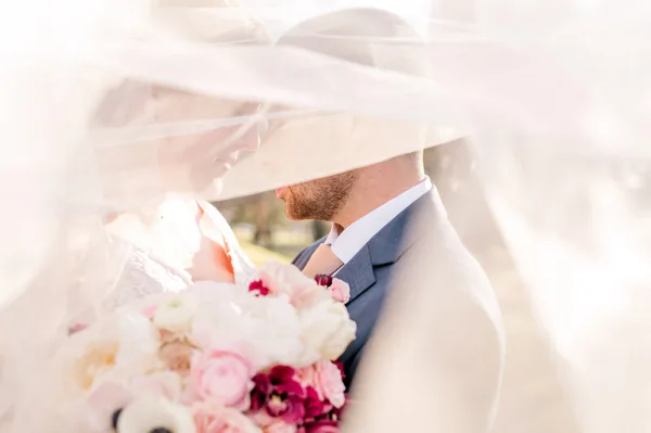 A bride and groom sharing a tender moment under a veil, with the bride holding a bouquet of pink and white flowers.