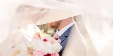 A bride and groom sharing a tender moment under a veil, with the bride holding a bouquet of pink and white flowers.