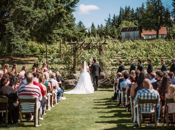 Outdoor wedding ceremony with bride and groom at a floral arch, surrounded by seated guests and a wedding party in a grassy field.