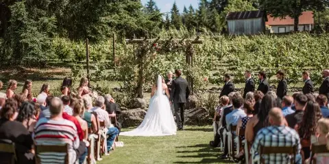 Outdoor wedding ceremony with bride and groom at a floral arch, surrounded by seated guests and a wedding party in a grassy field.