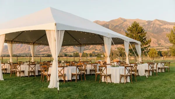 White event tent set up on a grassy field with round tables, chairs, and white linens, with mountains in the background.