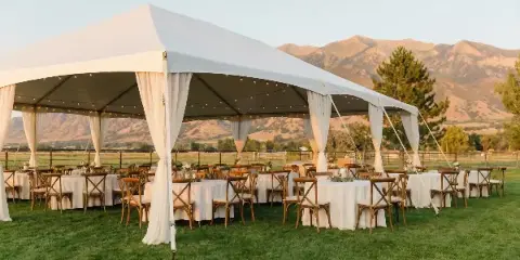 White event tent set up on a grassy field with round tables, chairs, and white linens, with mountains in the background.
